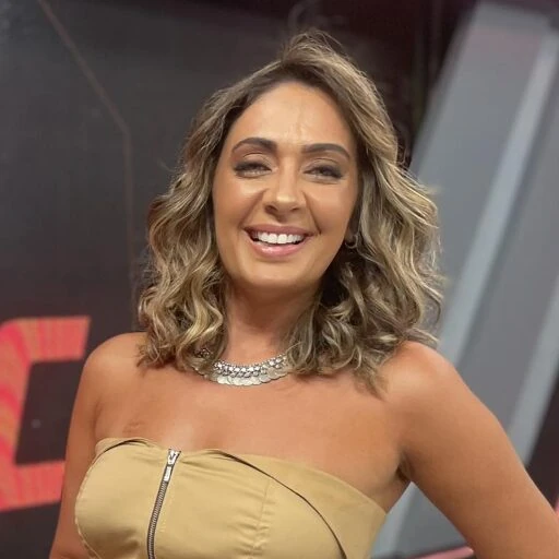 Mirelle Moschella sorrindo em cenário de TV, com colar e vestido dourado